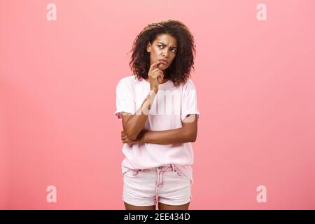 Suspect mécontent à la peau sombre jeune femme aux cheveux bouclés dans un t-shirt à la mode et short disant hmm touchant le menton avec le doigt se froissant et regardant Banque D'Images