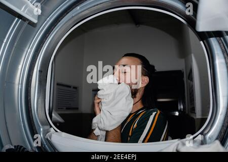 homme aux cheveux longs dans la salle à laver les vêtements sentant le doux parfum qui a été imprégné dans ses vêtements blancs, sèche-linge industriel. industriel wa Banque D'Images