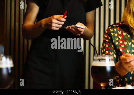 Serveuse prenant et commander de la femme blonde sur le chemisier vert dans le restaurant, café, pizzeria. Uniforme noir. Tenant le stylo rouge et le petit ordinateur portable. Bière activée Banque D'Images