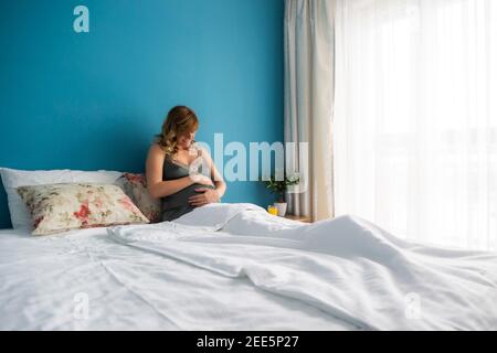 Une femme enceinte souriante en position semi-assise au lit avec ses mains autour de son estomac. Elle regarde son estomac. Banque D'Images