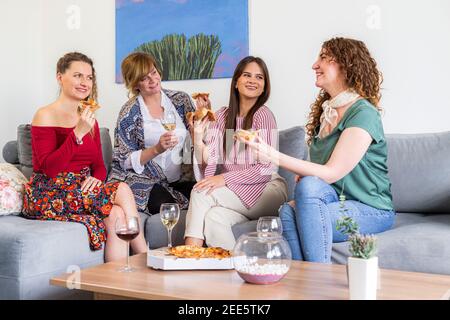 Quatre amies souriantes bavardent et s'asseyant sur le canapé dans le salon tout en mangeant de la pizza, il y a aussi des verres avec du vin sur la table. Banque D'Images