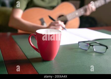 Blur femme musicienne avec guitare écrivant sur le livre au bureau. Mise au point sélective sur la tasse rouge. Banque D'Images