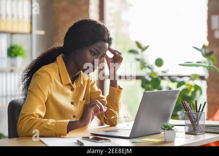 Photo portrait d'une femme afro-américaine qui touche la tête et qui travaille sur ordinateur portable dans un bureau moderne à l'intérieur Banque D'Images