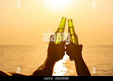 Couple mains tenant des bouteilles de bière claquant et célébrant en vacances à la plage en été coucher de soleil Banque D'Images