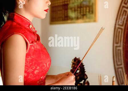 Femme asiatique en rouge qipao robe priant avec des bâtons d'encens Durant la nouvelle année chinoise ou lunaire Banque D'Images