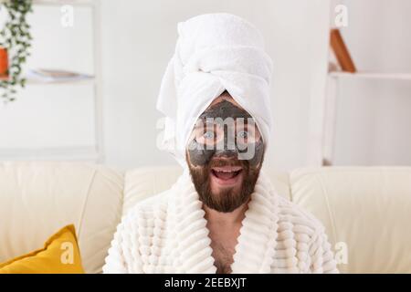 Homme barbu s'amusant avec un masque cosmétique sur son visage en argile noire. Hommes soins de la peau, humour et spa à la maison concept Banque D'Images