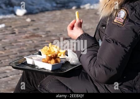 Une femme sous un manteau d'hiver épais mange du poisson et des frites assis à l'extérieur sur un banc dans une rue. Concentrez-vous sur l'épaule et le poisson Banque D'Images