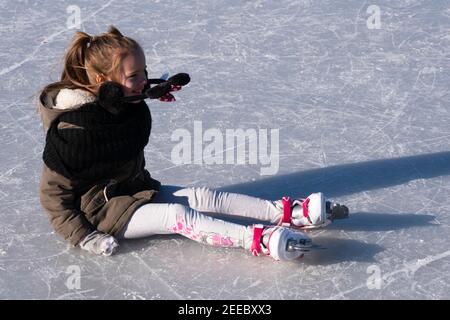 Petite fille, vêtue de vêtements d'hiver épais, s'assoit sur la glace, elle est juste tombée en patinage. Heureusement, elle peut rire à ce sujet. Vue latérale Banque D'Images