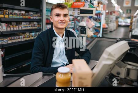 La caisse des jeunes hommes travaille à la caisse de l'épicerie. Homme assis derrière le comptoir de facturation, regardant la caméra et souriant. Banque D'Images