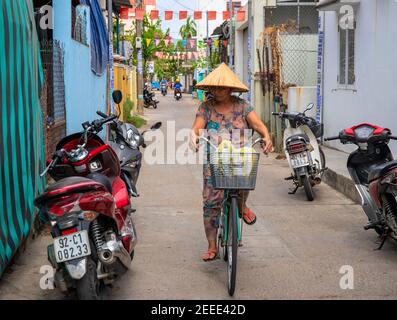 Hoi an, Vietnam - 28 juillet 2019: Femme dans chapeau vietnamien traditionnel à vélo dans la rue étroite. La vie quotidienne de la ville vietnamienne. Personne en conica Banque D'Images