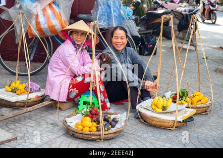 Hoi an, Vietnam - 28 juillet 2019: Femmes vendeur avec mât porteur assis dans la rue. Femmes vietnamiennes souriantes en chapeau conique vietnamien. Fruits tropicaux s Banque D'Images