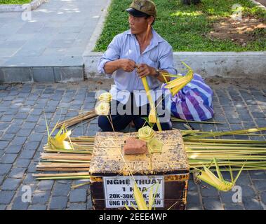 Hoi an, Vietnam - 28 juillet 2019 : homme faisant des fleurs à partir de feuilles de palmier. Vendeur de souvenirs dans un lieu touristique. Un meneur vietnamien au travail. Fleur tressée à l'herbe Banque D'Images