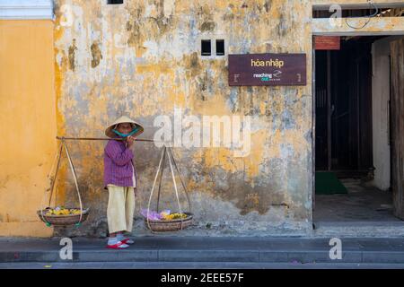 Hoi an, Vietnam - 28 juillet 2019: Vieille dame en chapeau vietnamien avec mât de transport, mur historique sur fond. Femme souriante en chapeau conique vietnamien. Tr Banque D'Images