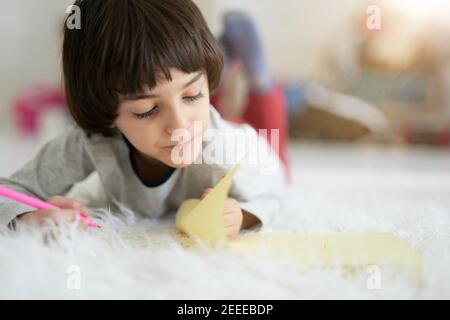 Gros plan de petit garçon latin regardant concentré tout en dessinant des images avec des crayons colorés, couché sur le sol. Enfant impliqué dans l'activité créative. Enseignement à distance pour les enfants Banque D'Images