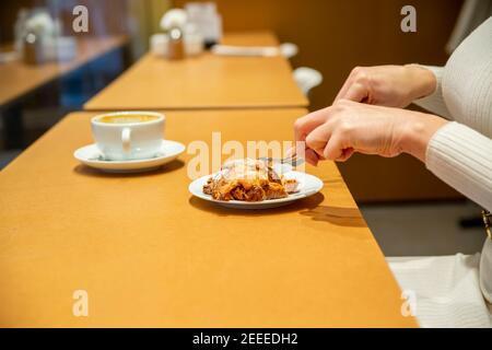 une femme coupe un croissant et boit du café à une table dans un café. pas de visage Banque D'Images