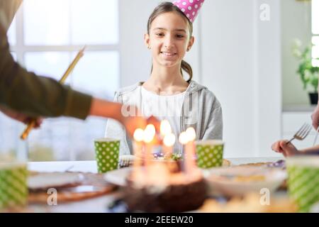 Bonne adolescente latine en chapeau d'anniversaire souriant et regardant le gâteau d'anniversaire tout en célébrant l'anniversaire avec sa famille à la maison. Enfants, concept de fête Banque D'Images