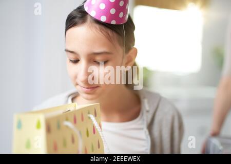 Curieuse jeune fille hispanique vérifiant, regardant à l'intérieur du sac cadeau, tout en recevant des cadeaux, célébrant l'anniversaire à la maison. Célébration, concept d'enfance Banque D'Images