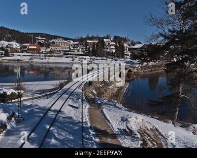 Voies ferrées sur la rive du lac gelé Schluchsee avec pont ferroviaire et village en arrière-plan le jour ensoleillé d'hiver avec ciel bleu. Banque D'Images