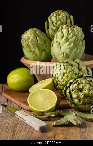 Vue d'artichauts verts dans un bol, de limes sur une table et un couteau, sur une table en bois, avec mise au point sélective, fond noir, vertical Banque D'Images