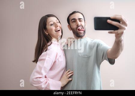 Couple prise de selfie, photo, chat vidéo, photographie, faire des visages, plaisantant, parlant en ligne tenant le téléphone. Décontracté pull à capuche rose pastel. Fond vide Banque D'Images