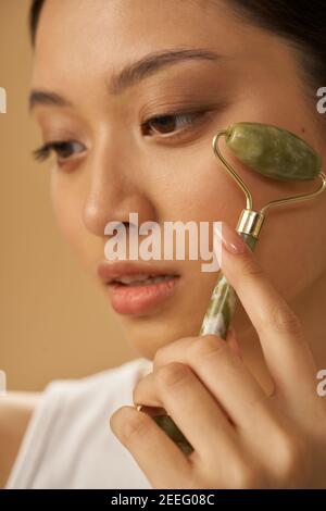 Gros plan sur le visage d'une jeune femme attirante regardant de côté et utilisant un rouleau de jade pour masser son visage, posant isolé sur fond beige. Concept de soin de la peau. Tir vertical Banque D'Images