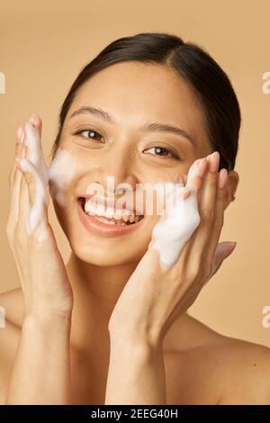 Portrait de beauté de la jeune femme joyeuse souriant à l'appareil photo tout en appliquant une mousse douce nettoyant pour le visage isolé sur fond beige. Produits de beauté et concept de soins de la peau Banque D'Images