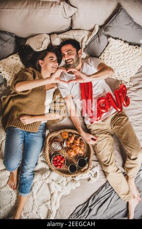 Jeune couple heureux allongé sur des couvertures pieds nus dans le lit, prenant le petit déjeuner avec des croissants et des fraises et faisant le coeur des doigts avec signe d'amour, vue de dessus. Saint Valentin concept de jour Banque D'Images