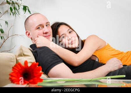 Double portrait inclinable d'un jeune couple hétérosexuel qui s'embrasse un canapé à la maison devant un mur lumineux regarder le camérier Banque D'Images