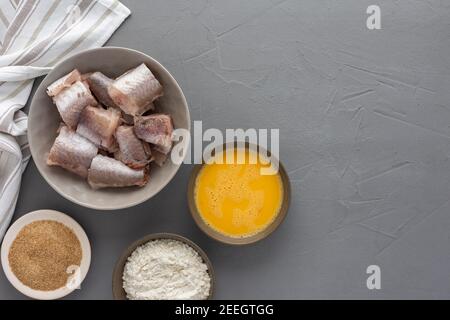 Dans un bol, des morceaux de merlu et des produits pour la cuisine frite, des œufs, de la farine et de la chapelure ont été présentés avec une serviette de cuisine sur fond gris Banque D'Images