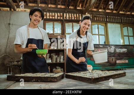 homme et femme souriants portant un tablier regardant l'appareil photo tout en remuant la pâte à gâteau dans un bol près du moule à gâteau sur la table. Banque D'Images