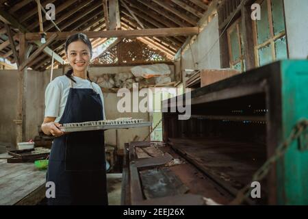 une fille portant un tablier sourit à l'appareil photo alors qu'elle apporte une plaque de cuisson remplie de pâte à biscuits à mettre dans le four déjà éclairé avec la cuisine en arrière-plan. Banque D'Images