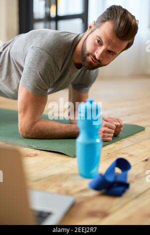 Homme sportif barbu regardant l'entraînement en ligne sur ordinateur portable et de la performance planche d'avant-bras à la maison Banque D'Images