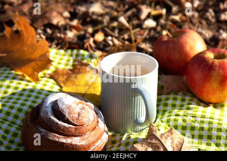 L'automne encore la vie. Pommes rouges mûres, une tasse de café chaud avec du lait dans une tasse bleue, un petit pain à la cannelle doux sur une nappe à carreaux verts Banque D'Images