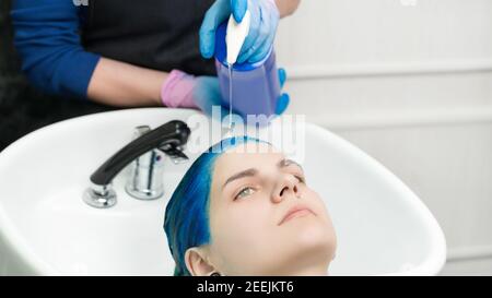 Le coiffeur passe les mains en gants pour comprimer le shampooing du tube dans la tête jeune femme avec des cheveux bleus tout en se lavant les cheveux en spécial salon de beauté avec lavabo et douche Banque D'Images