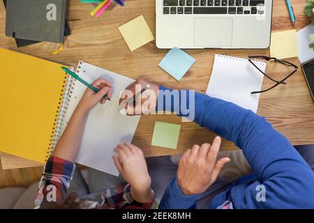 Vue de dessus du père latin attentionné aidant son fils à faire ses devoirs, expliquant la tâche, prenant des notes tout en étant assis à la maison. Apprentissage à distance, famille, paternité concept Banque D'Images