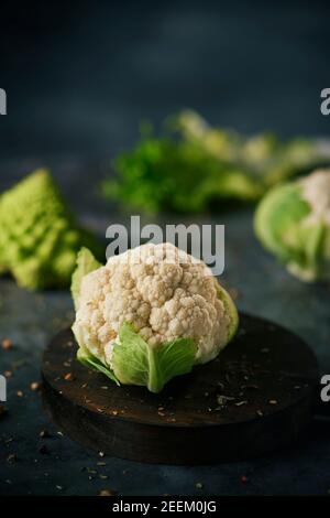 une tête de chou-fleur brute dans une assiette en bois, placée sur une surface en pierre sombre parsemée de différentes graines et épices et un bébé brocoli romanesco Banque D'Images