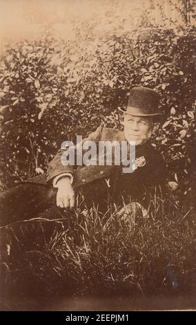 Carte postale photographique vintage du début du XXe siècle montrant un homme âgé vêtu d'un costume et portant un chapeau. L'homme est posé sur l'herbe à côté de quelques buissons. Banque D'Images