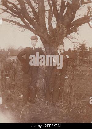 Vintage 1906 Edwardian carte postale photographique montrant deux garçons d'école debout devant un arbre. Banque D'Images