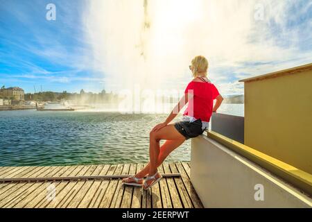 Une jeune fille touristique assise sur une jetée en bois, regardant la fontaine Jet d'eau qui projette son jet d'eau de 140 mètres de haut.l'attraction la plus célèbre et Banque D'Images