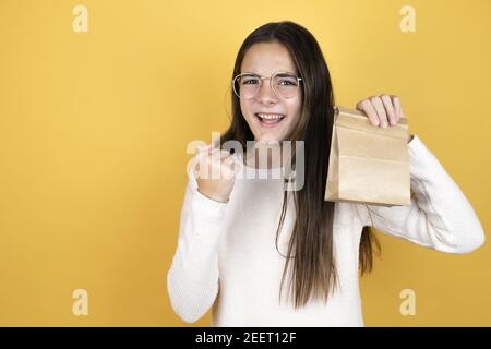 Belle enfant fille portant des vêtements décontractés tenant un sac en papier très heureux et excités faisant le geste gagnant avec les bras relevés, souriant et criant f Banque D'Images