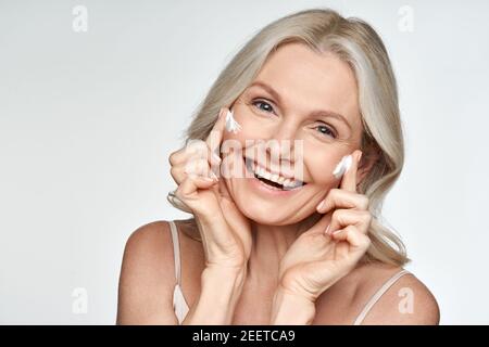 Bonne femme d'âge moyen appliquant de la crème sur la peau du visage isolée sur fond blanc. Banque D'Images