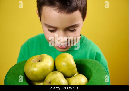 Le petit garçon aime le parfum des pommes vertes dans une casquette irlandaise verte. Portrait sur fond jaune. Saint Patrick Banque D'Images