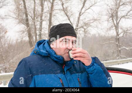 Gros homme d'âge moyen dans une veste chaude et un chapeau boit une boisson chaude en plein air en hiver. Reposez-vous au froid sur le chemin. Banque D'Images