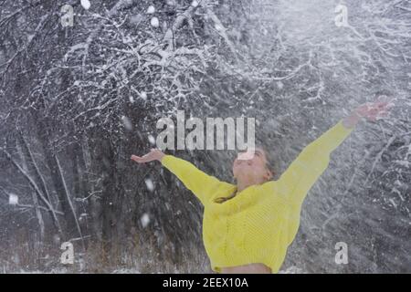 Joyeuse jeune femme jette une poignée de neige poudreuse fraîche haut dans l'air le jour d'hiver. Lady dans un chandail jaune vif dans la forêt givrée pour la marche. Banque D'Images