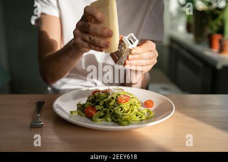 Gros plan des mains de femme râpant le parmesan dans les pâtes avec sauce pesto, tomates cerises fraîches. Banque D'Images