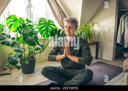 Jeune homme assis sur un tapis tout en faisant le yoga position Namaste dans la chambre à coucher avec des plantes. Cours de yoga en ligne. Un style de vie sain. Accueil en ligne Banque D'Images