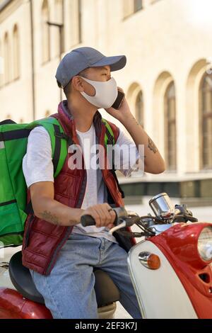 Photo verticale d'un jeune messager asiatique avec thermo sac portant un masque de protection pour le visage parlant par téléphone portable avec client lors de la livraison de nourriture par scooter automobile pendant la quarantaine du coronavirus Banque D'Images