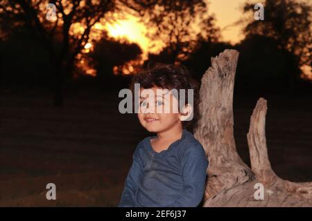 Un beau petit enfant indien, assis sur un bois sec dans le jardin au coucher du soleil, enfant indien souriant regardant la caméra. Concept pour Sweet c Banque D'Images