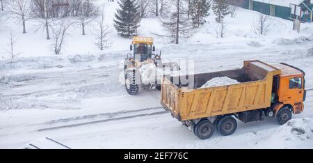 Le gros tracteur jaune nettoie la neige de la route et la charge dans le camion. Nettoyage et nettoyage des routes de la ville de la neige en hiver Banque D'Images