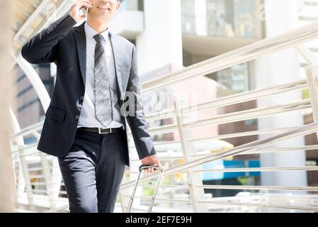 Homme d'affaires qui appelle sur un téléphone portable tout en tirant ses bagages et en marchant dans un passage couvert extérieur Banque D'Images